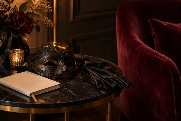 Editorial still life of a black mask, silk gloves and a notebook beside a burgundy velvet chair